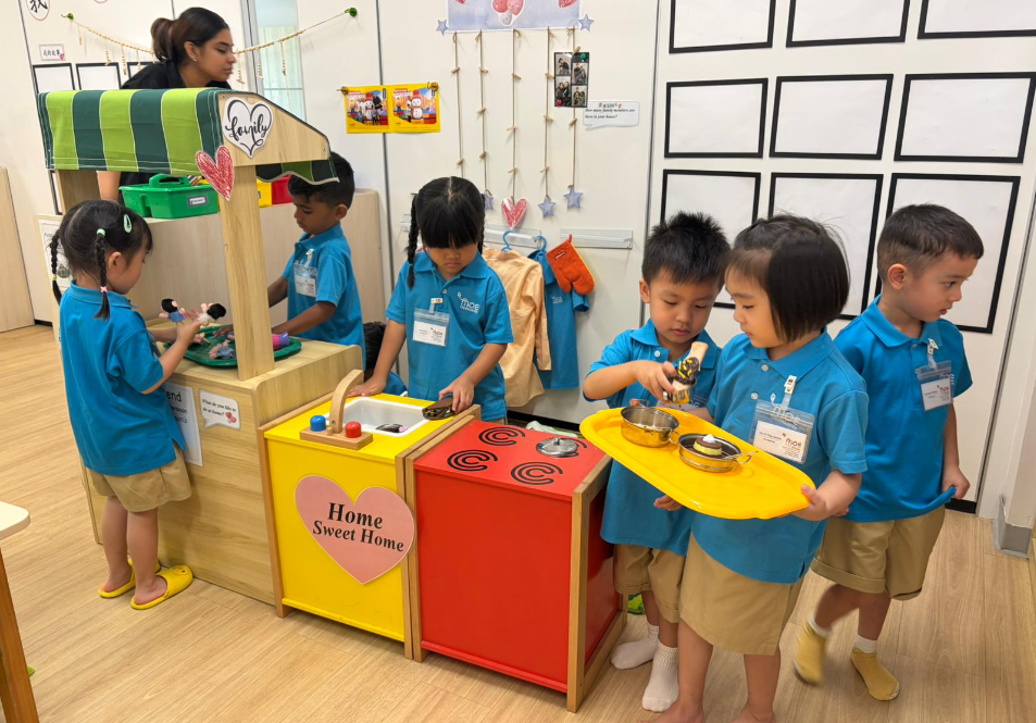 Children in blue uniforms play kitchen, using toy stove and sink while a teacher watches in a classroom.